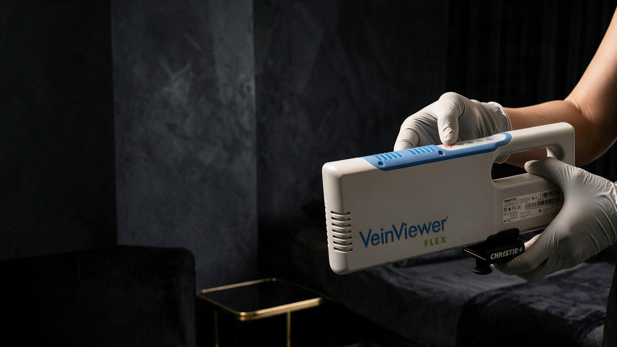 Gloved hands holding the VeinViewer Flex imaging device in a dark treatment room
