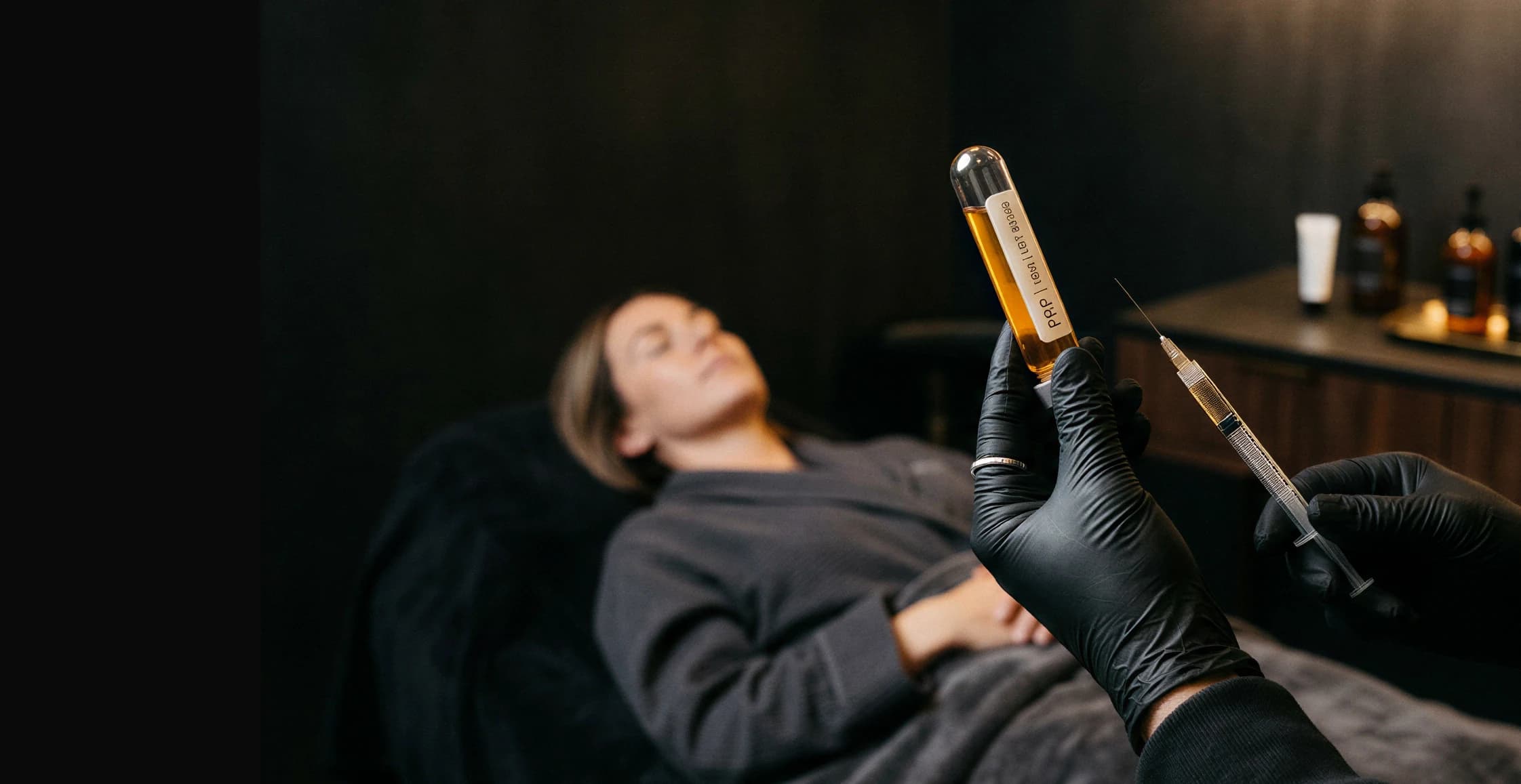 A clinician in black gloves holding a vial of platelet-rich plasma and a syringe beside a resting patient